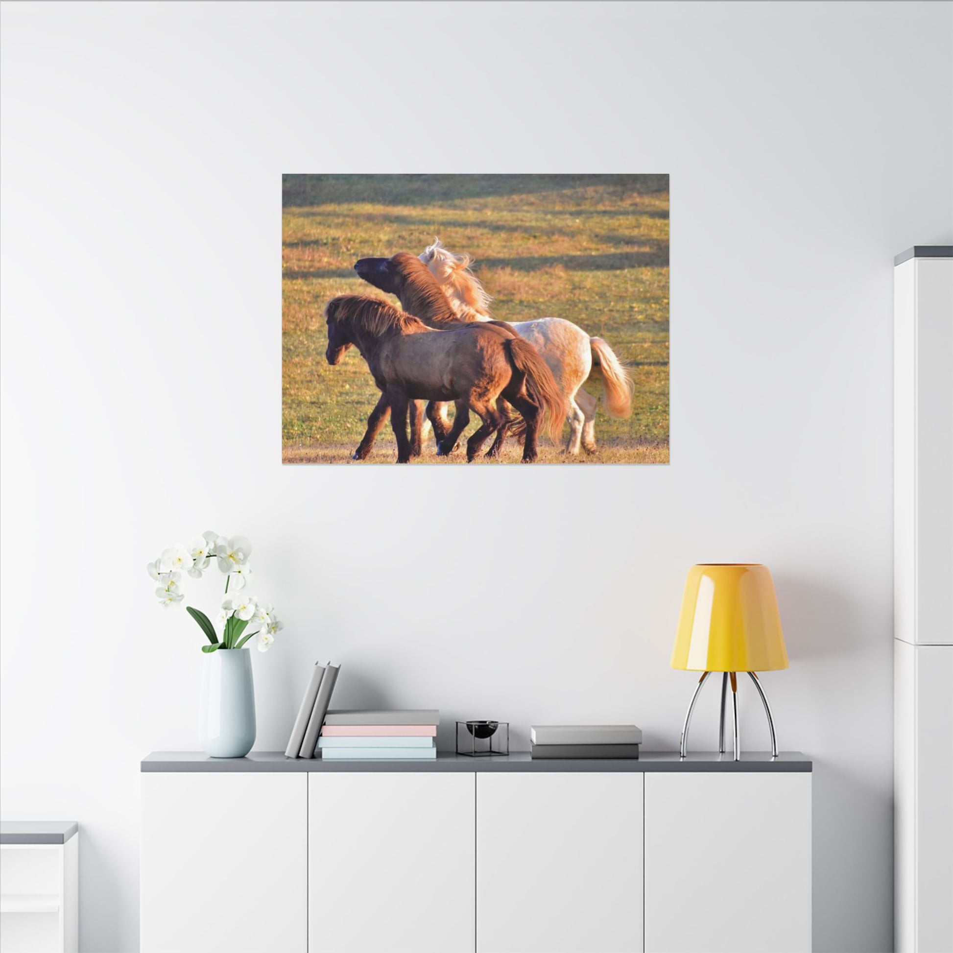 Three horses in a field depicted on a wall art piece above a white cabinet with a yellow lamp.